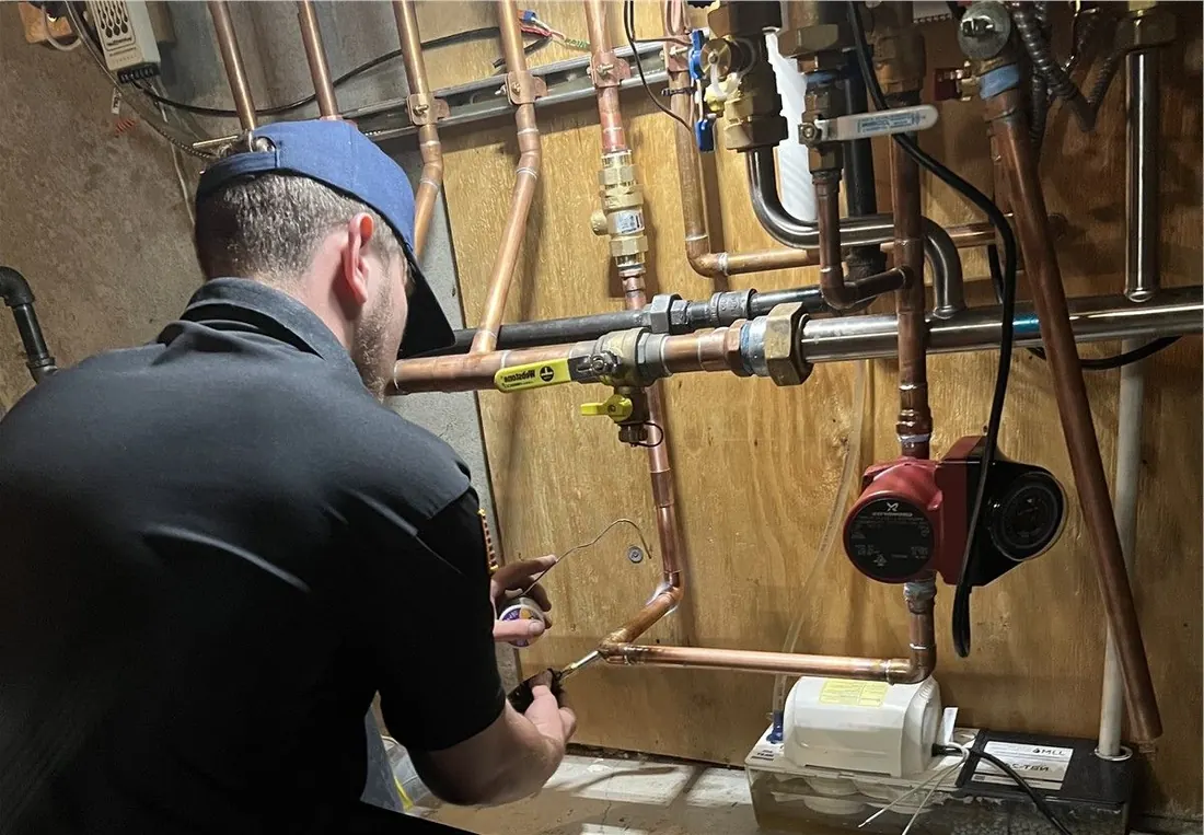 Plumber working on copper pipes and valves in a utility room. He wears a blue cap and black shirt.