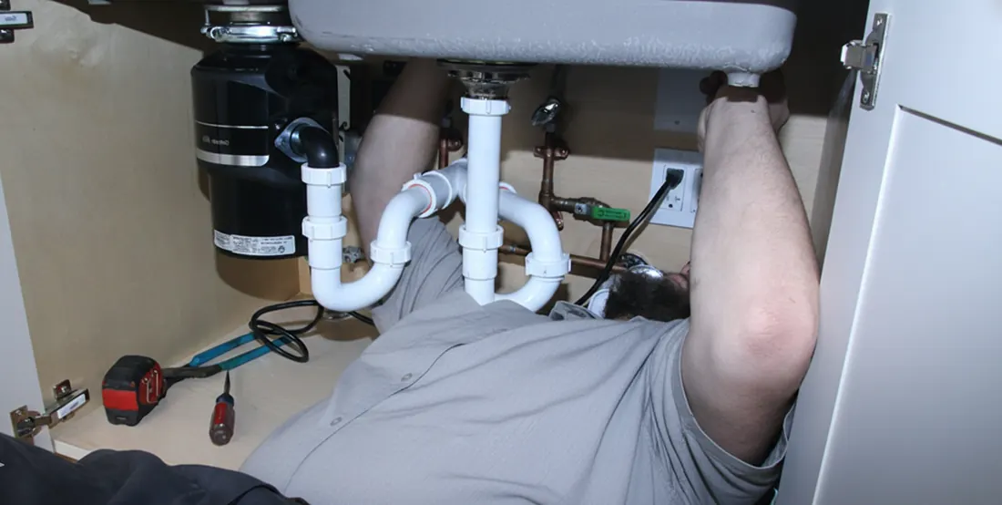 A person under a sink is working on plumbing. A garbage disposal, pipes, and tools are visible.